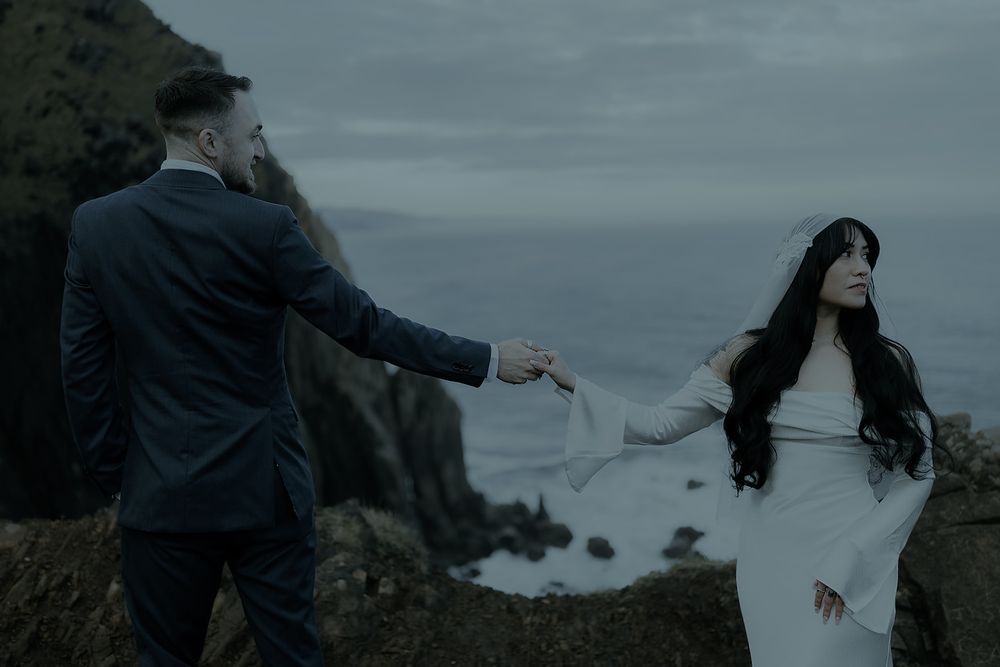 Couple in wedding attire holding hands on dramatic coastal cliffs with moody ocean waves in background.