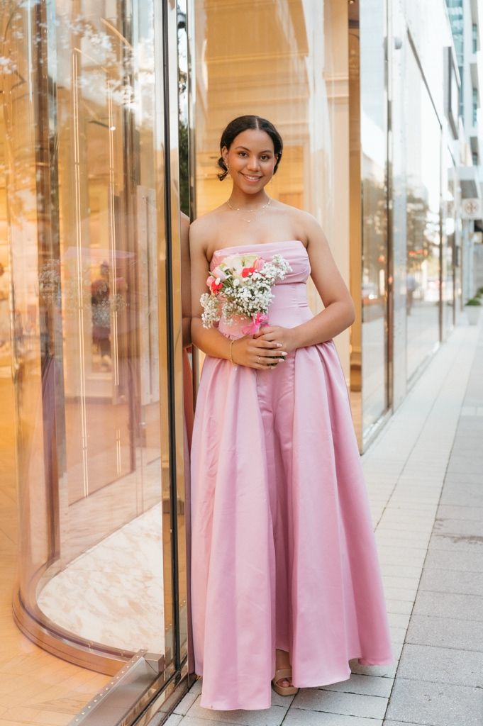 Young woman in strapless pink gown holding bouquet outside luxury store.