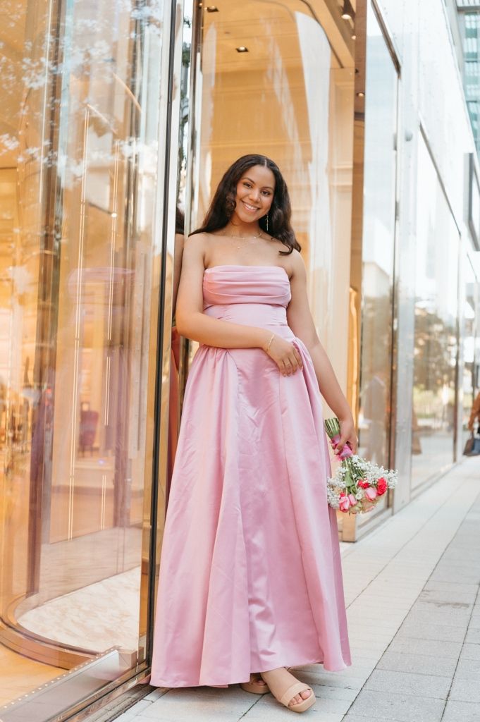 Young woman in elegant pink strapless ball gown holding flowers, posing outside luxury retail store.
