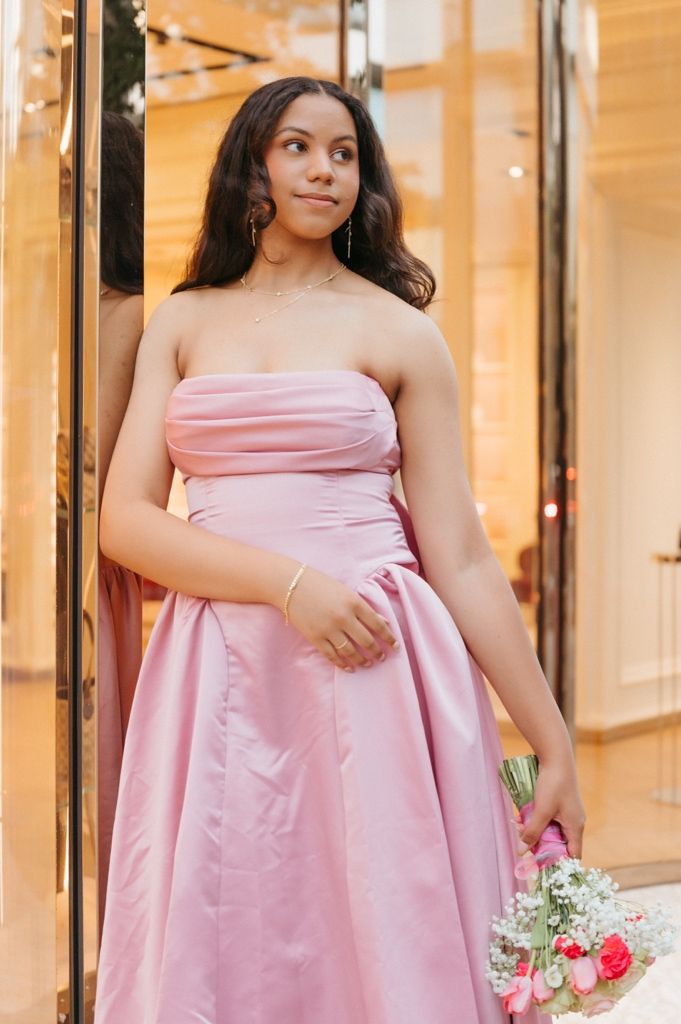 Young woman in a strapless pink ball gown holding a floral bouquet, posing elegantly near a glass storefront.