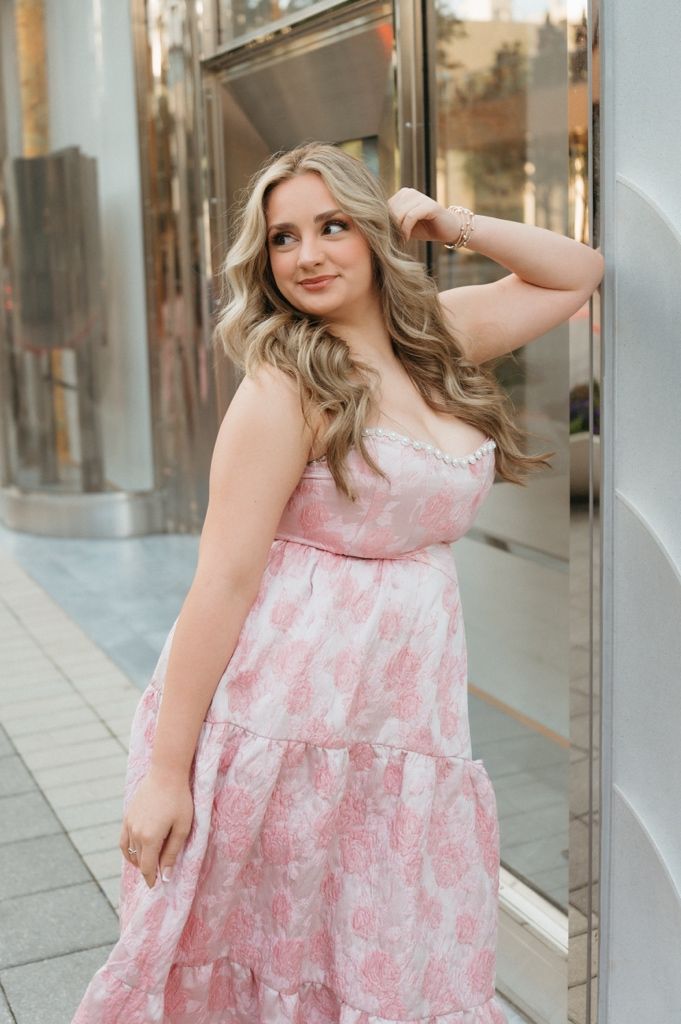 Young woman in pink floral tiered dress posing outdoors, leaning against a mirrored wall with wavy blonde hair.