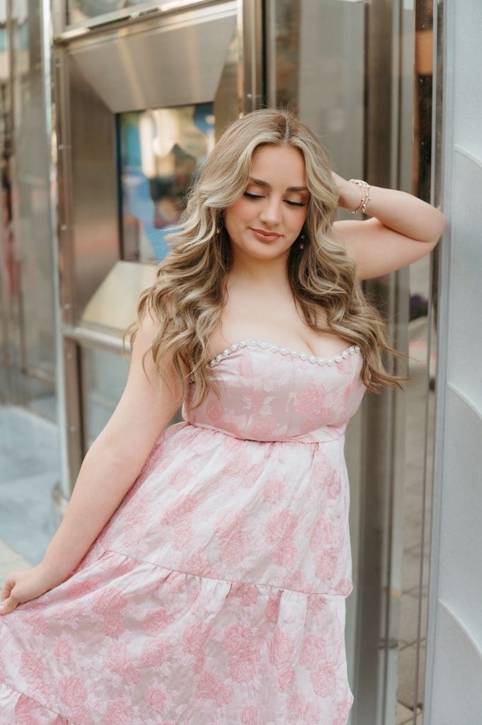 Young woman with long blonde wavy hair poses in a pink floral tiered dress outdoors.