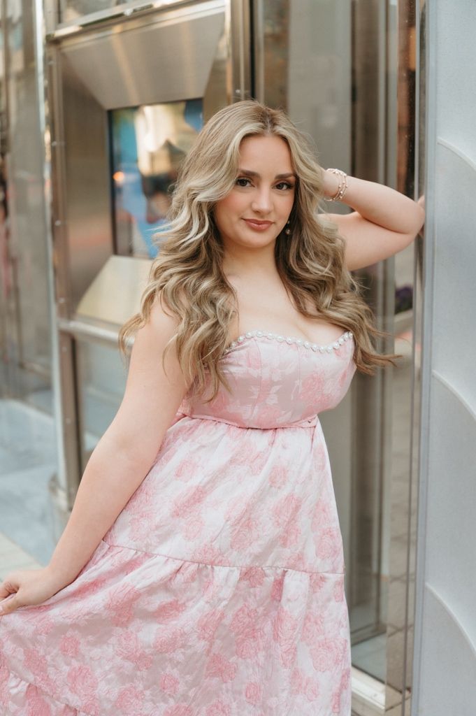 Young woman in pink floral strapless tiered maxi dress poses gracefully with hand in hair near silver door.