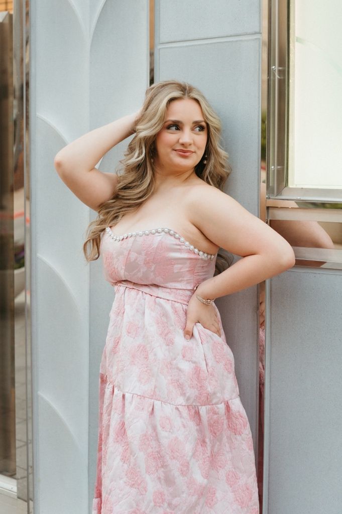 Young woman in a strapless pink floral maxi dress poses gracefully against a gray wall outdoors.