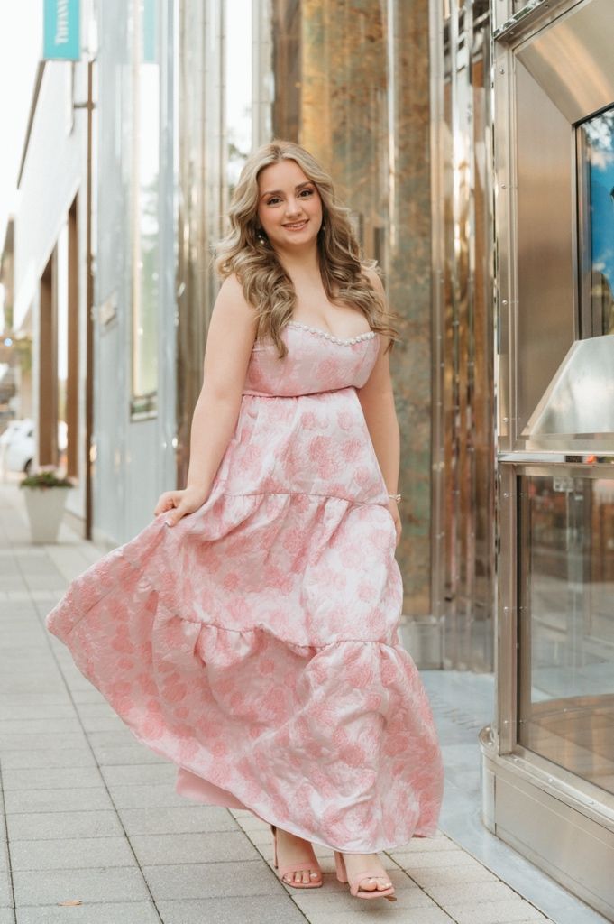 Young woman in pink floral strapless tiered maxi dress posing on urban sidewalk outside luxury store.