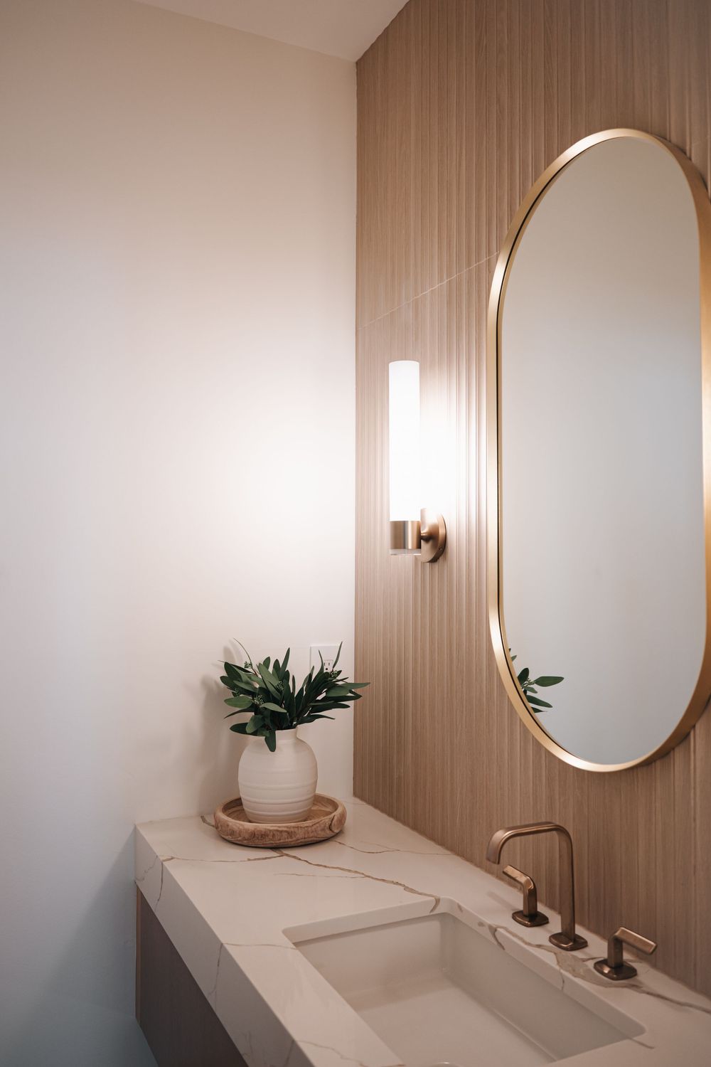 Modern bathroom with oval gold mirror, wood slat wall, marble vanity, brass faucet, and white ceramic vase with greenery.