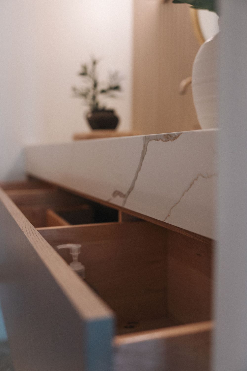 Marble countertop bathroom vanity with open wooden drawer, decorative plant and mirror in background.