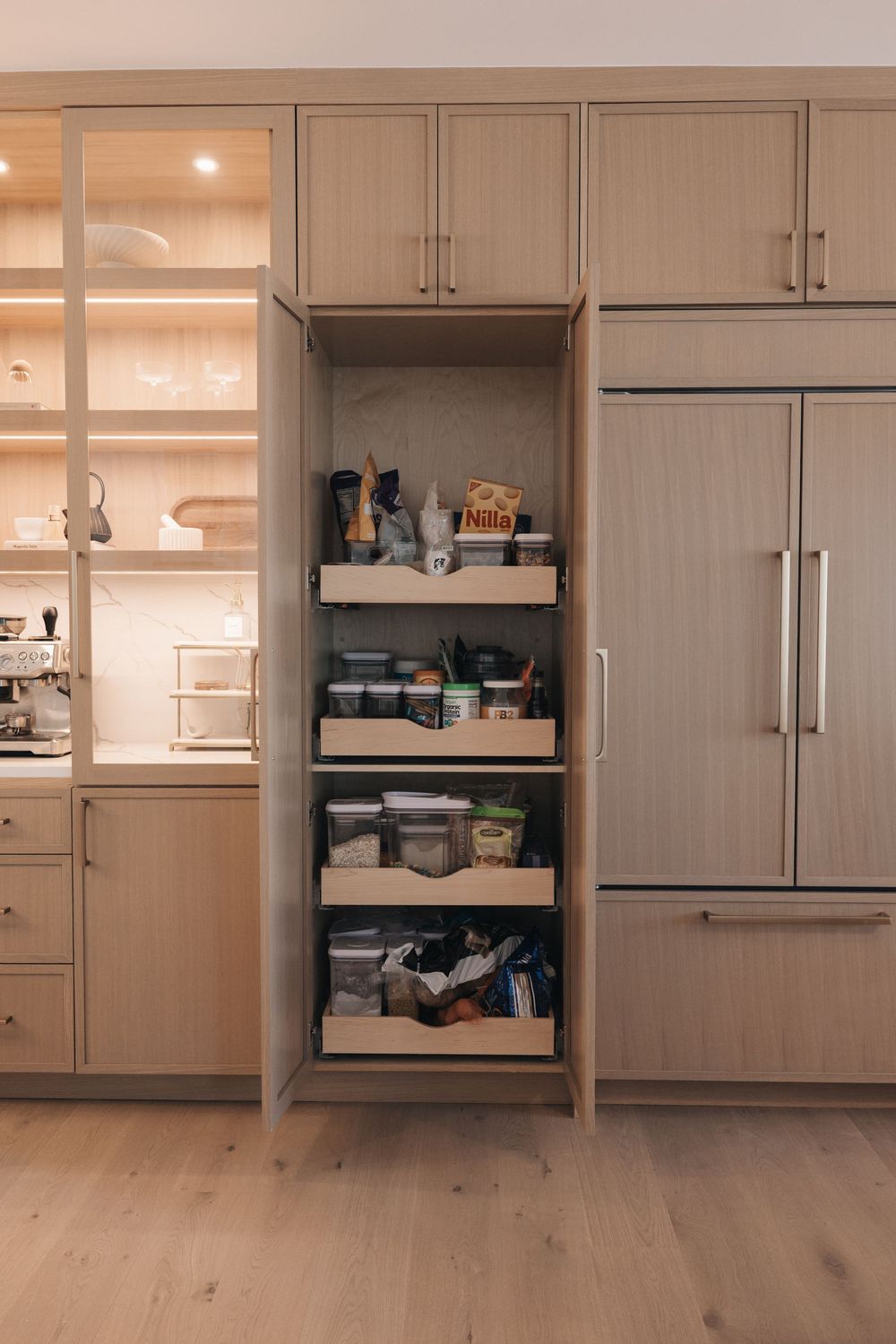 Modern kitchen pantry cabinet with pull-out shelves storing food items, flanked by glass-front cabinets and integrated refrigerator.