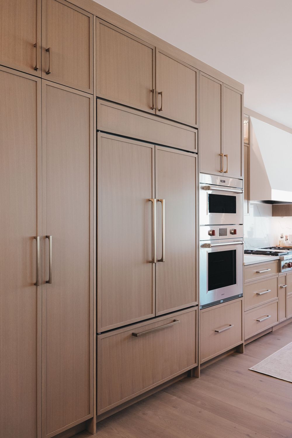 Modern kitchen with integrated panel-ready refrigerator, double wall ovens, and light wood cabinetry with brass hardware.