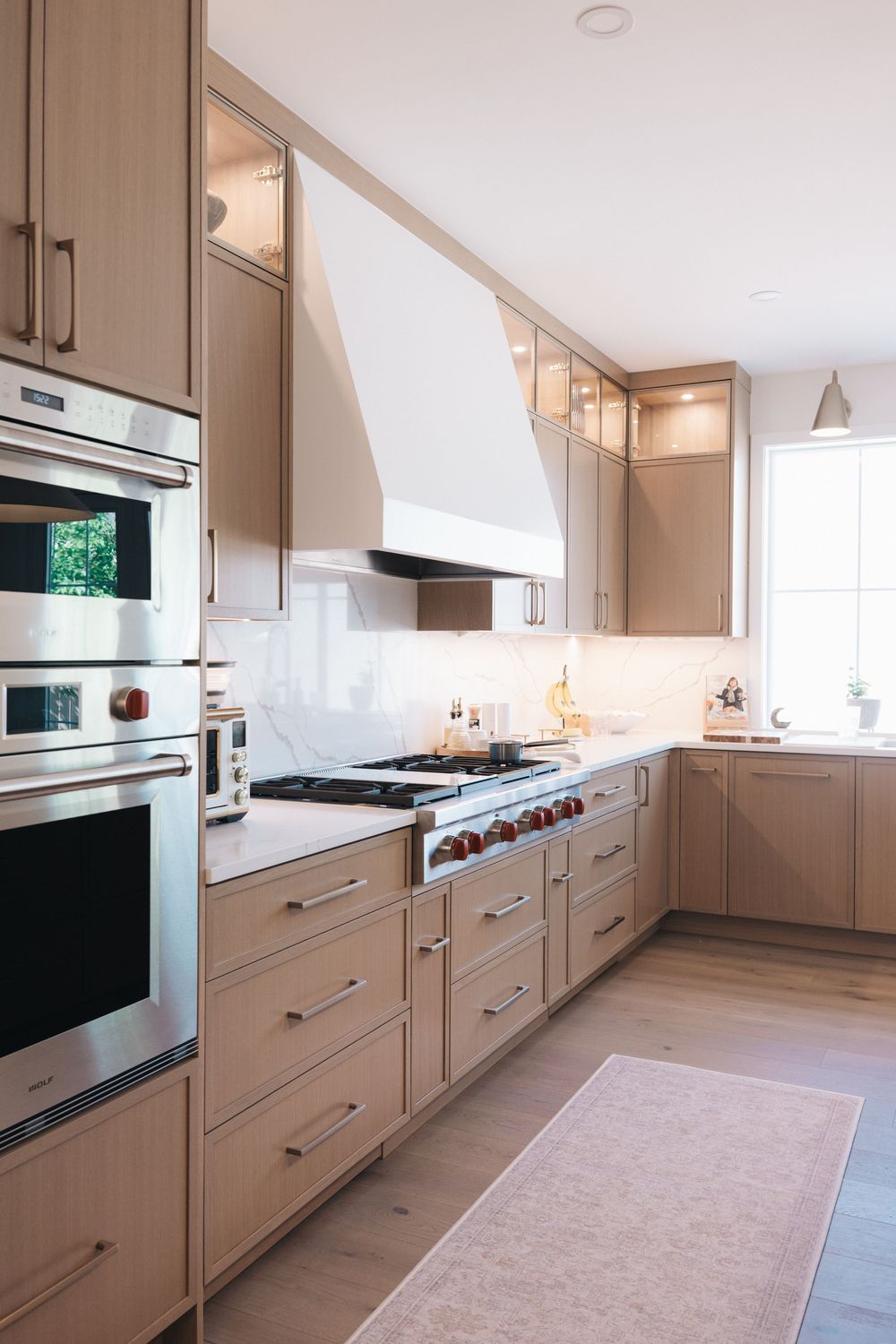 Modern kitchen with light wood cabinets, stainless steel appliances, white range hood, marble backsplash, and hardwood floors.
