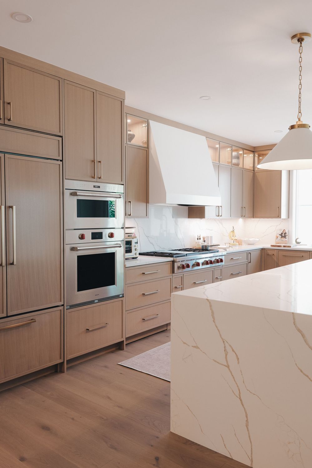 Modern kitchen with light wood cabinets, marble island, stainless steel appliances, white range hood, and gold pendant light.