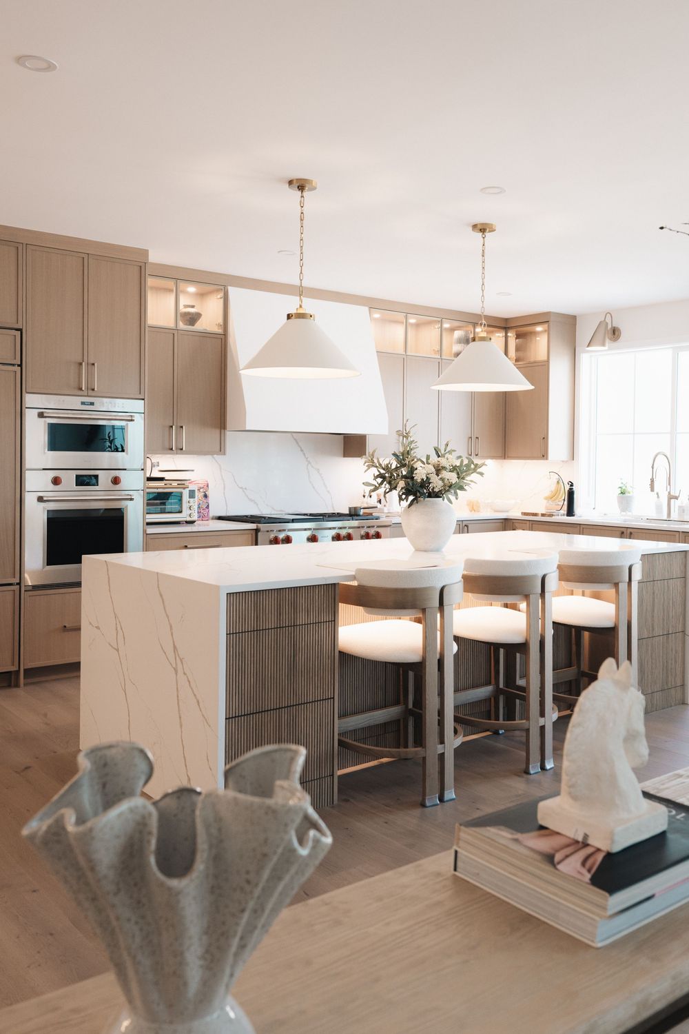 Modern kitchen with marble island, taupe cabinets, white pendant lights, bar stools, and stainless steel appliances on light hardwood floors