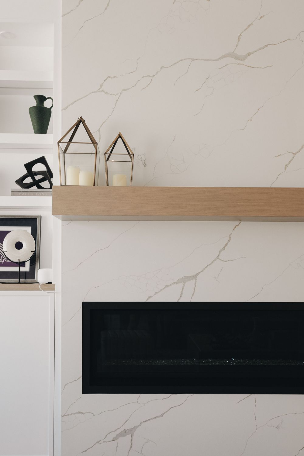 Modern linear fireplace with white marble surround, wood mantel, geometric gold lanterns, and built-in shelving with decor.