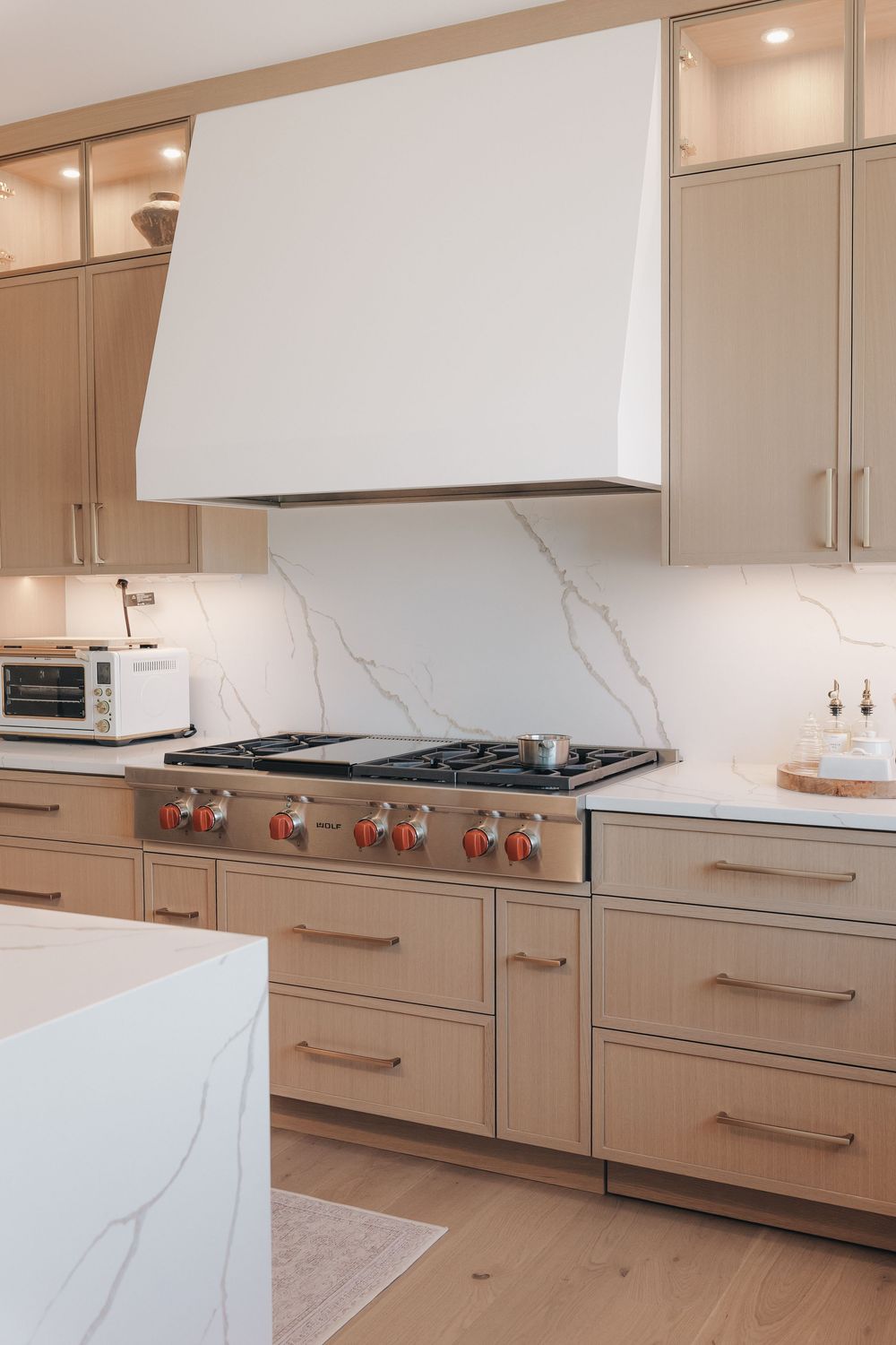 Modern kitchen with light wood cabinets, marble backsplash, professional gas range, and white hood vent.