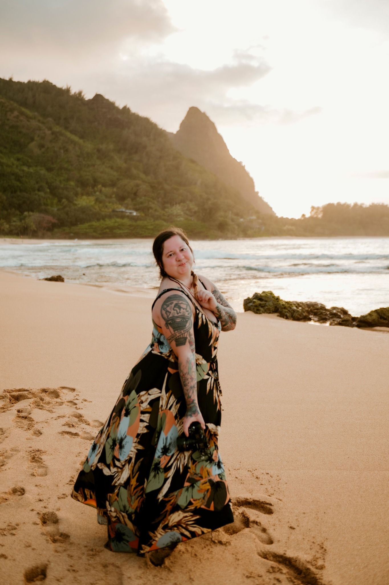Woman in floral maxi dress with tattoos poses on sandy Hawaiian beach at golden sunset with lush mountains backdrop.