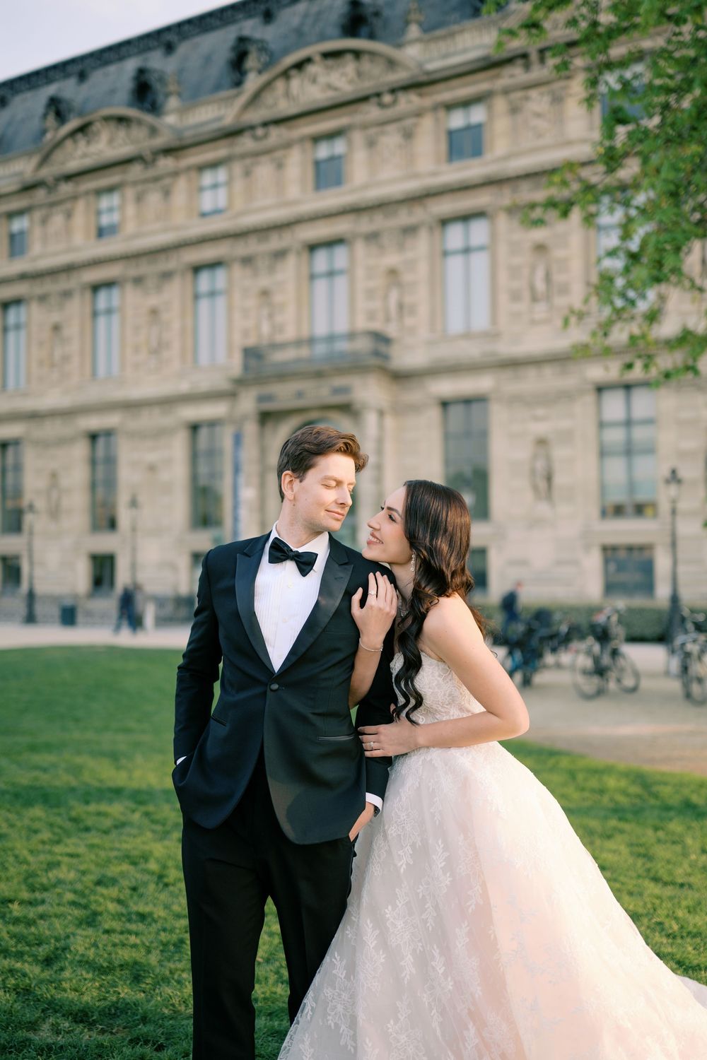 Elegant bride and groom posing in front of a grand European building, bride in lace gown, groom in black tuxedo.