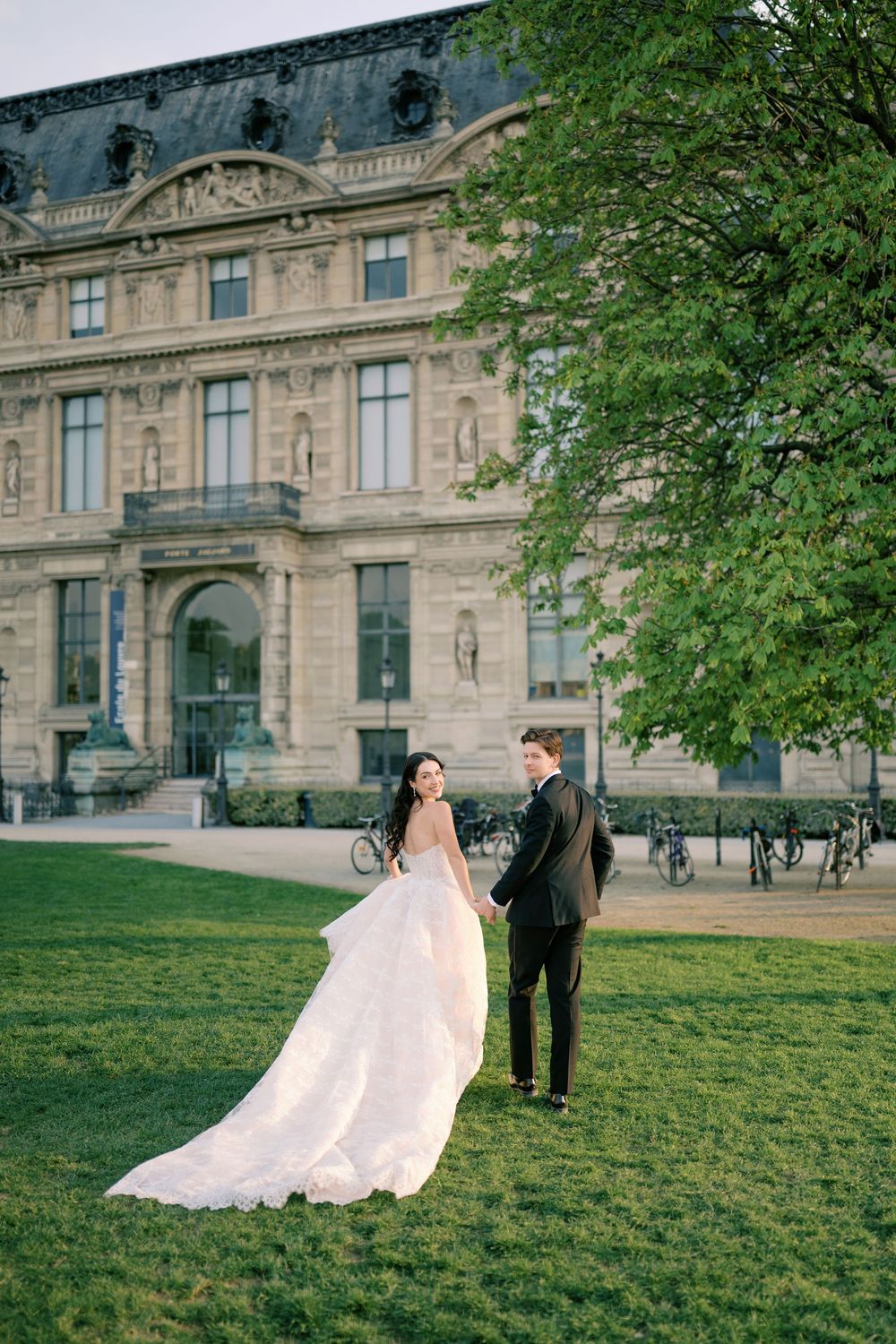 Bride and groom holding hands on green lawn before grand Parisian building, bride in flowing white gown.
