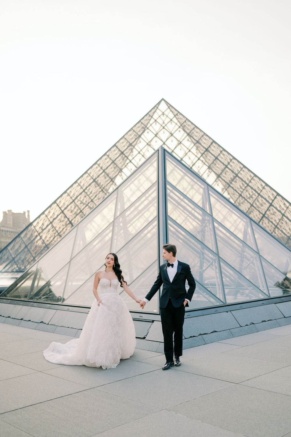 Bride and groom holding hands in front of the Louvre glass pyramid in Paris, wearing wedding gown and tuxedo.