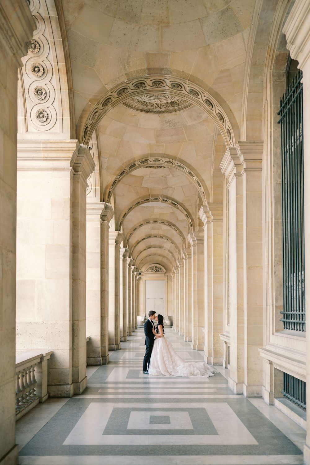 Bride and groom embracing under elegant stone arched colonnade in a grand classical European building.