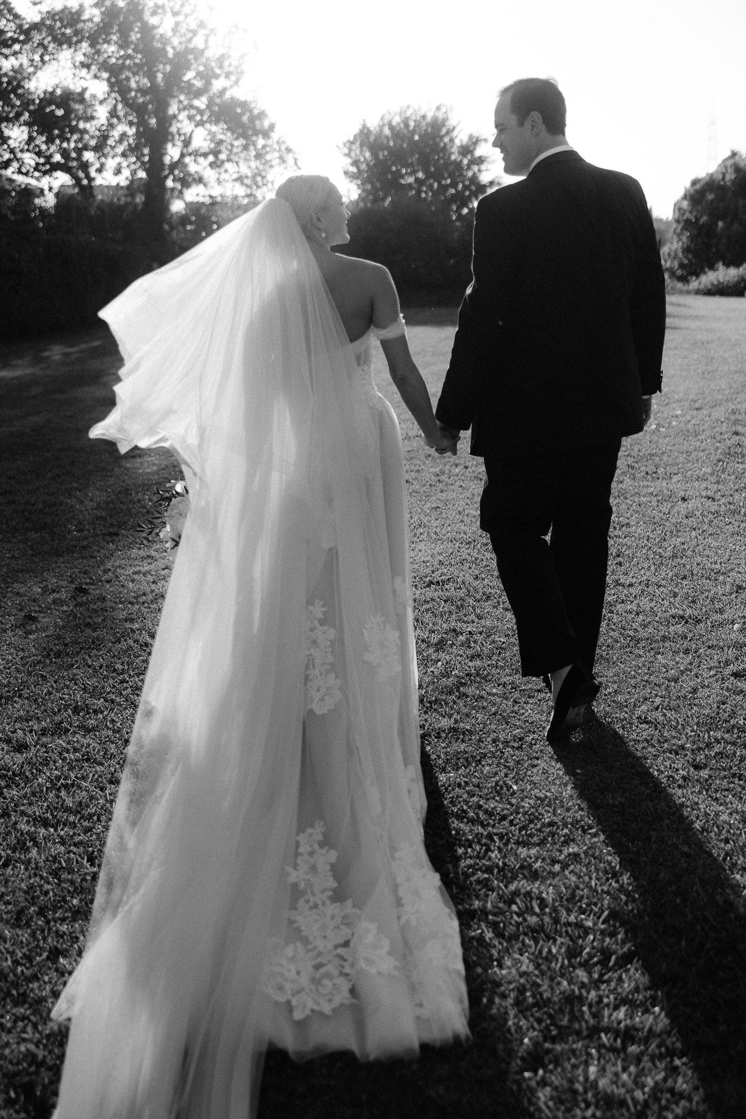 Black and white photo of bride and groom holding hands walking outdoors, veil flowing in wind.