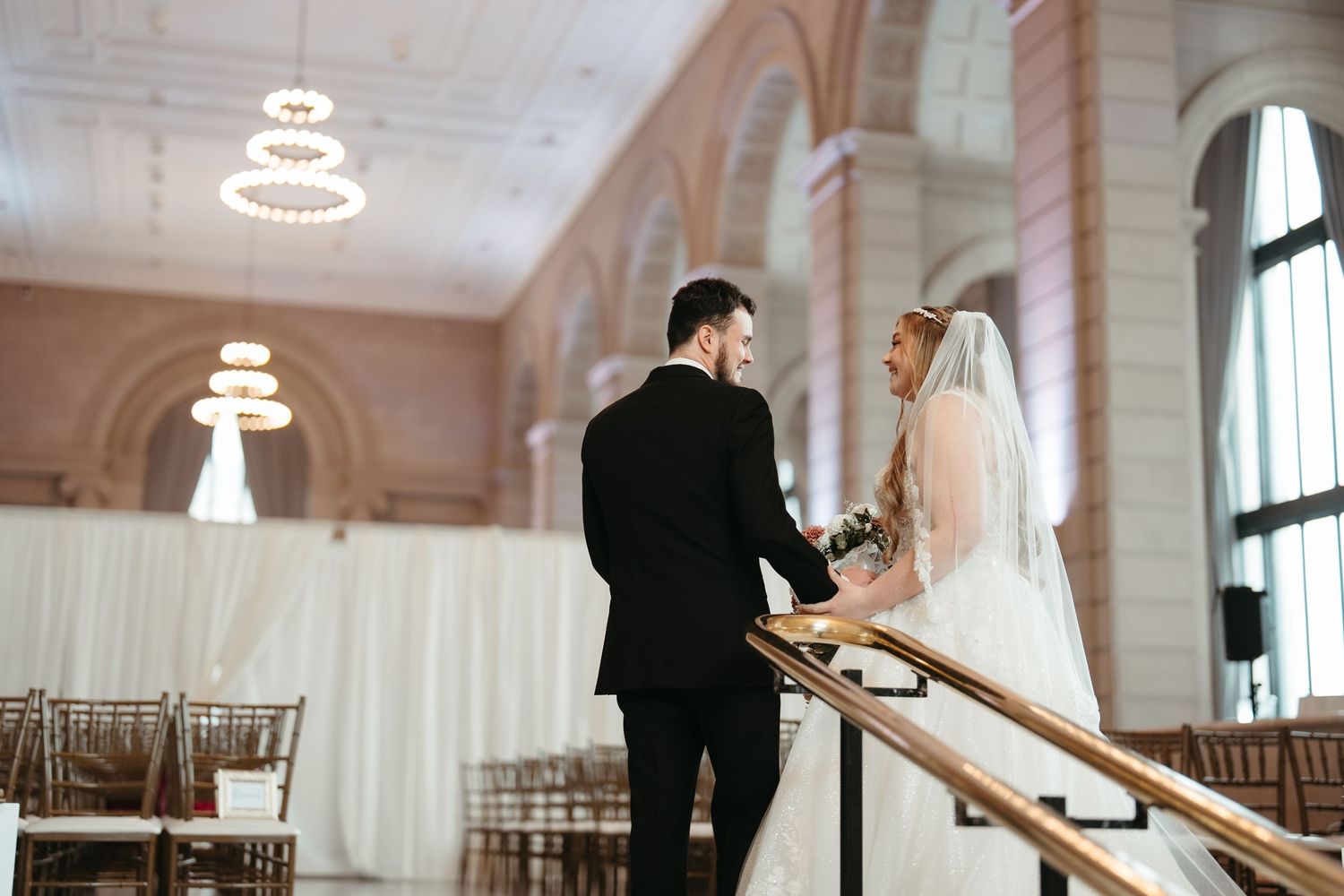 Bride and groom holding hands on elegant staircase in grand wedding venue with arched ceilings and chandeliers.