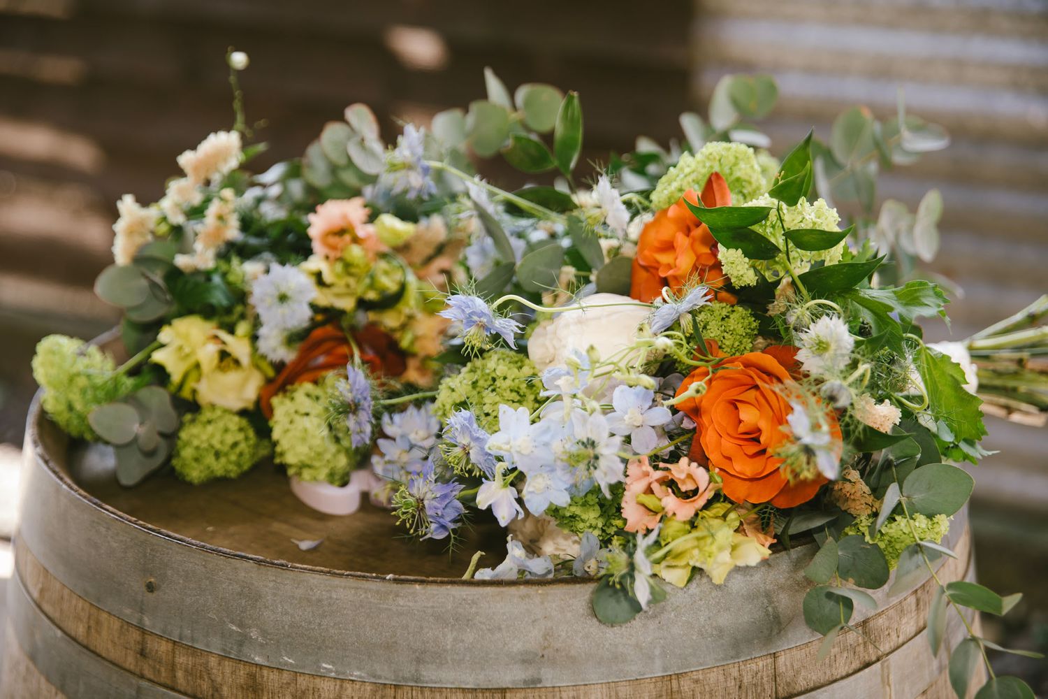 Colorful wedding bouquets with orange ranunculus, blue hydrangeas, and eucalyptus displayed on a rustic wooden barrel.