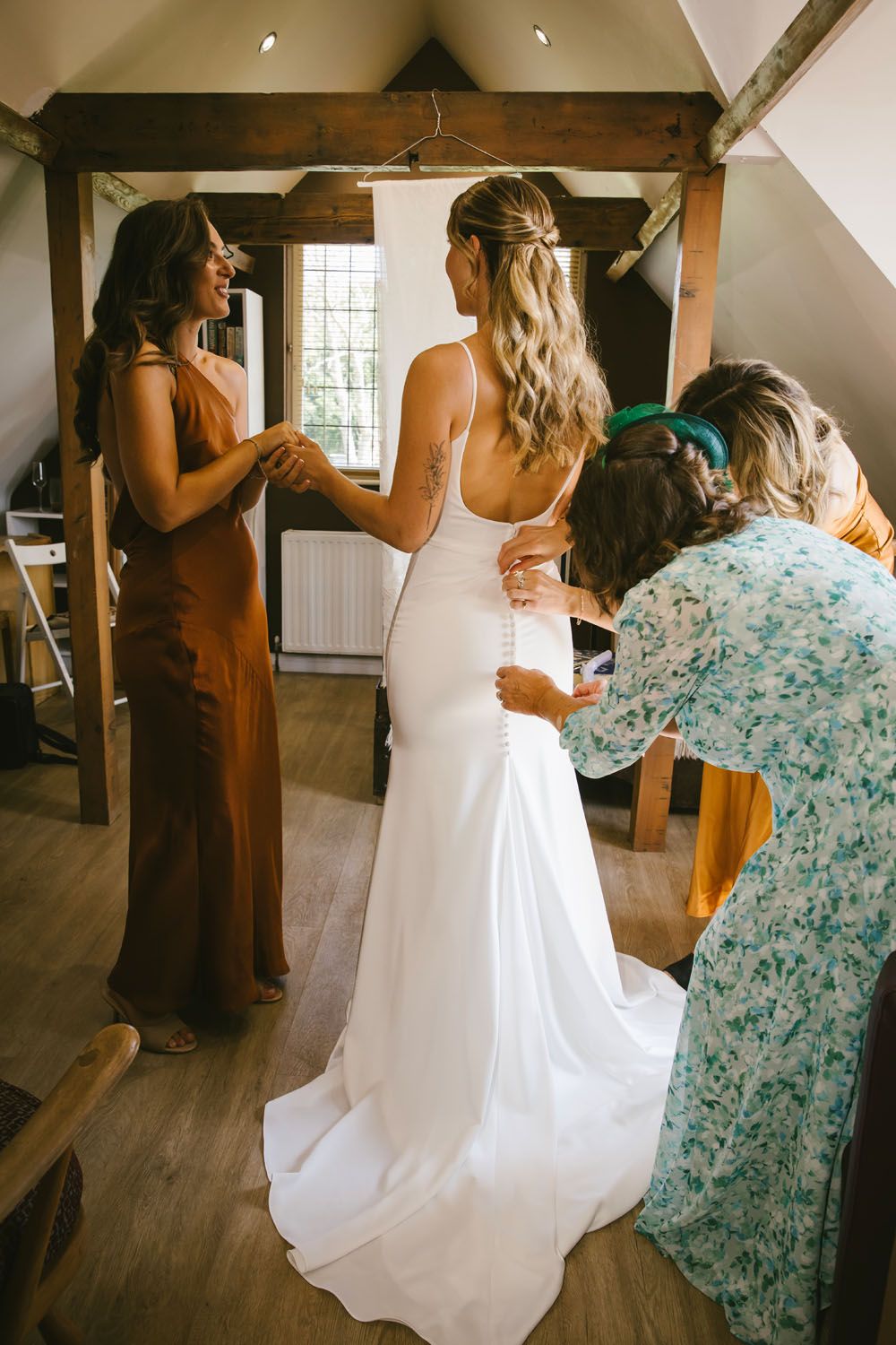 Bride in white gown being helped into dress by bridesmaids in rustic wooden room on wedding day.