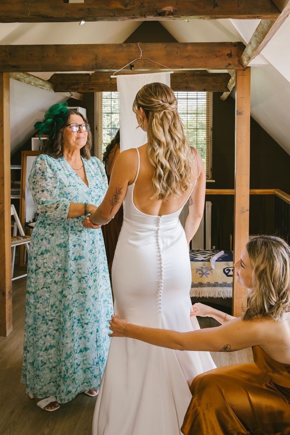 Bride in white gown getting dressed, assisted by bridesmaid in gold dress, while woman in floral dress watches in rustic venue.