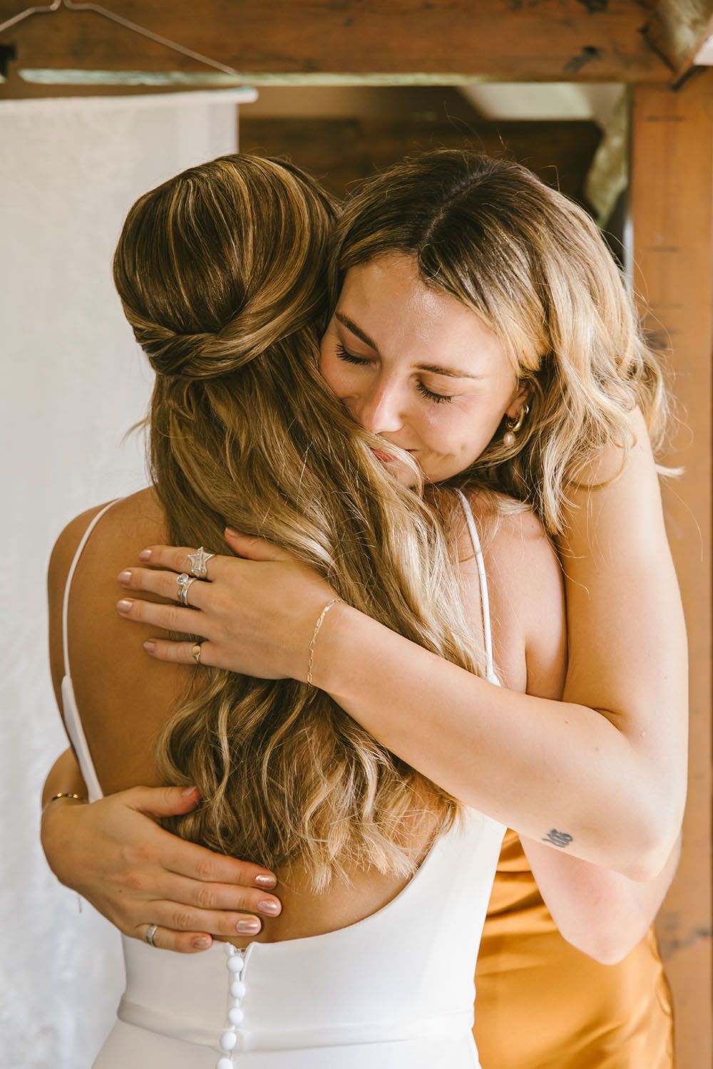 Two women sharing an emotional hug, one in a white bridal gown with elegant updo hairstyle at a wedding celebration.