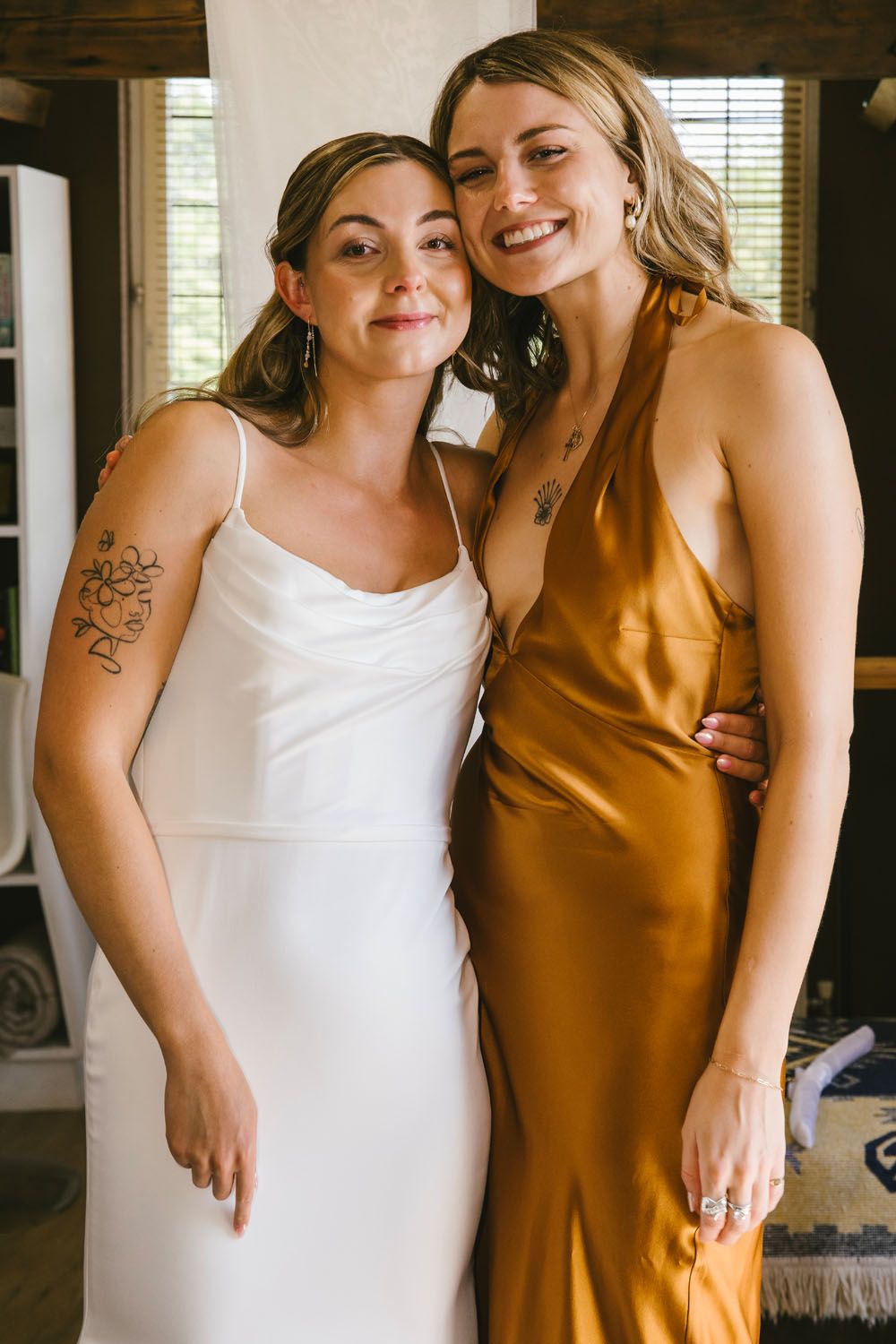 A bride in a white slip dress poses with a smiling bridesmaid in a golden satin gown, both showing arm tattoos.