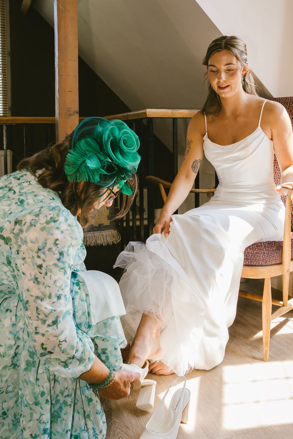 A smiling bride in a white dress sits as a woman in a green floral hat helps her put on her wedding shoes.