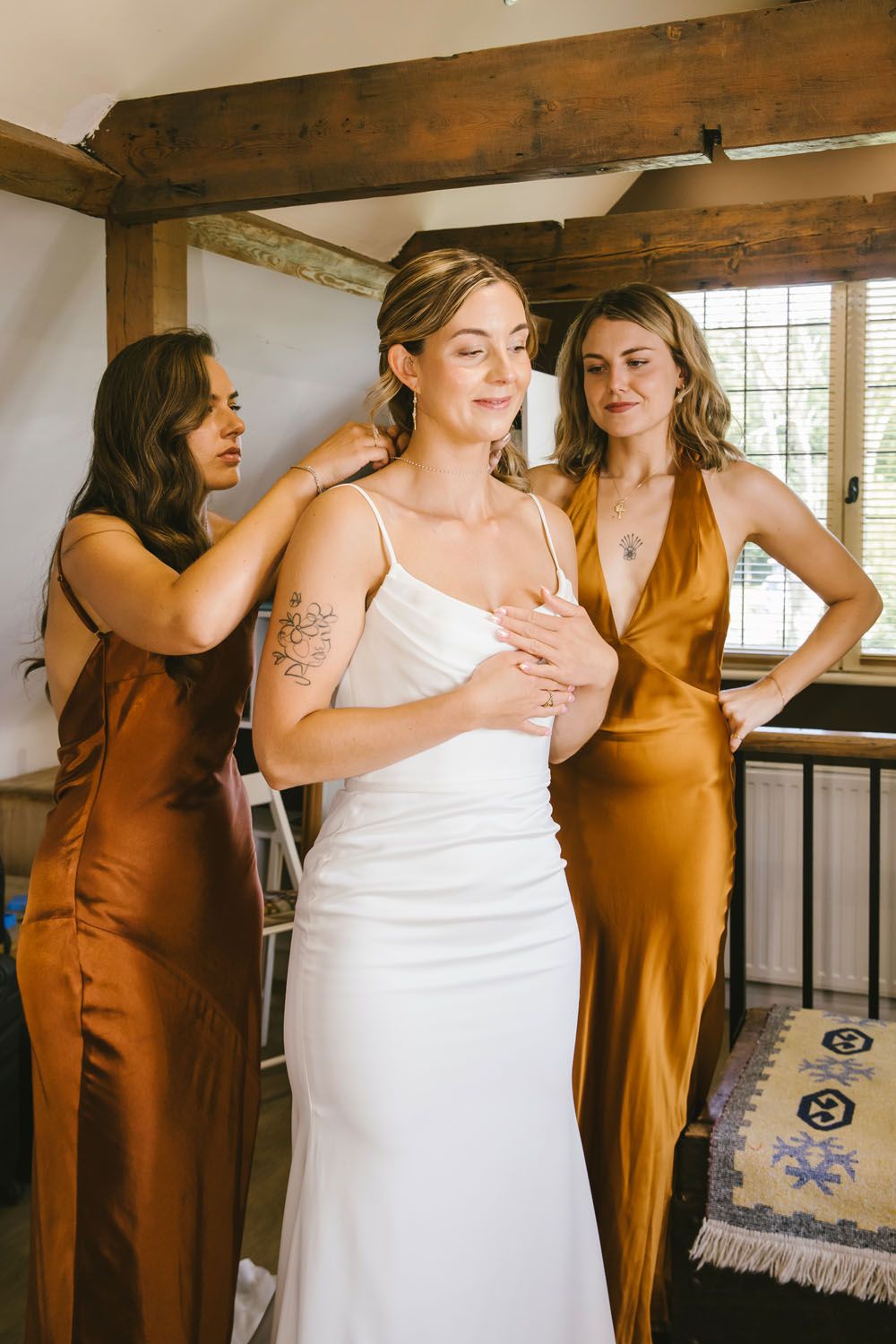 Bride in white gown assisted by two bridesmaids in gold satin dresses during wedding preparations indoors.