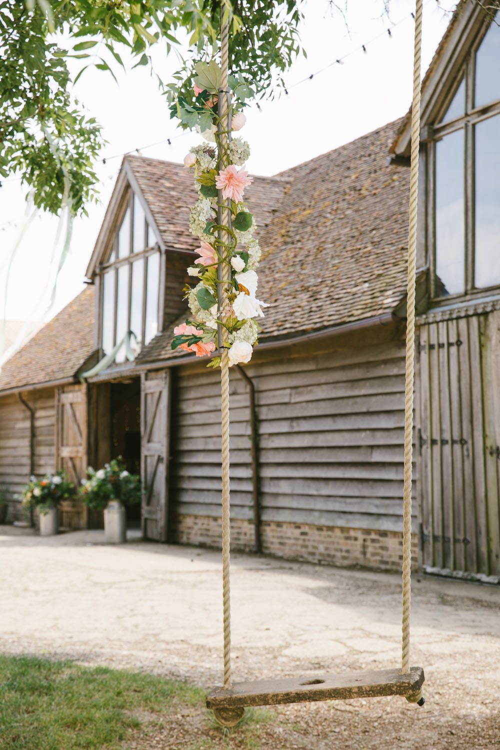 Rustic wooden swing adorned with pink and white flowers hanging by rope outside a charming barn venue.