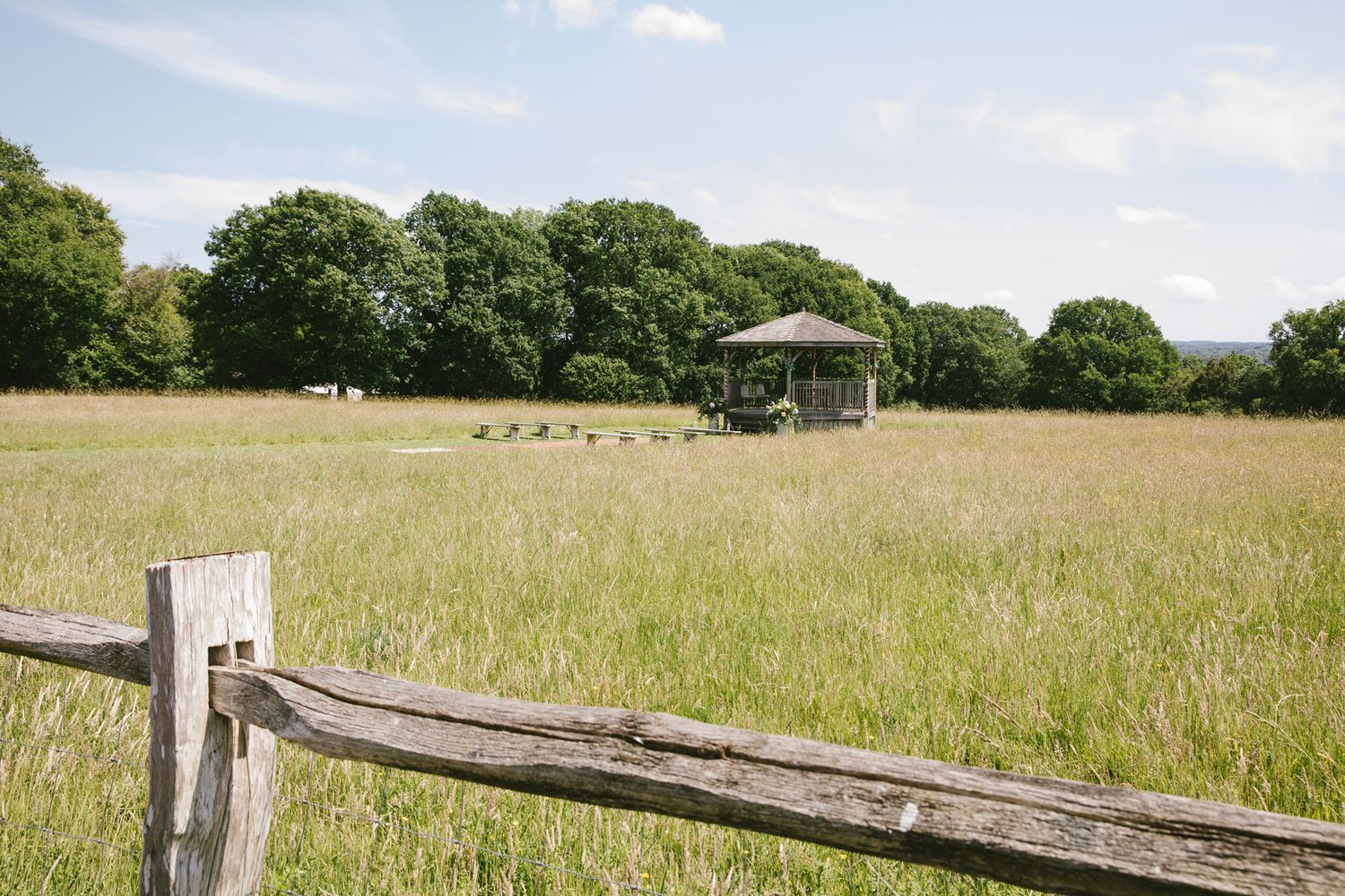 A rustic wooden fence borders a lush green meadow with a gazebo and trees in the background under a partly cloudy sky.