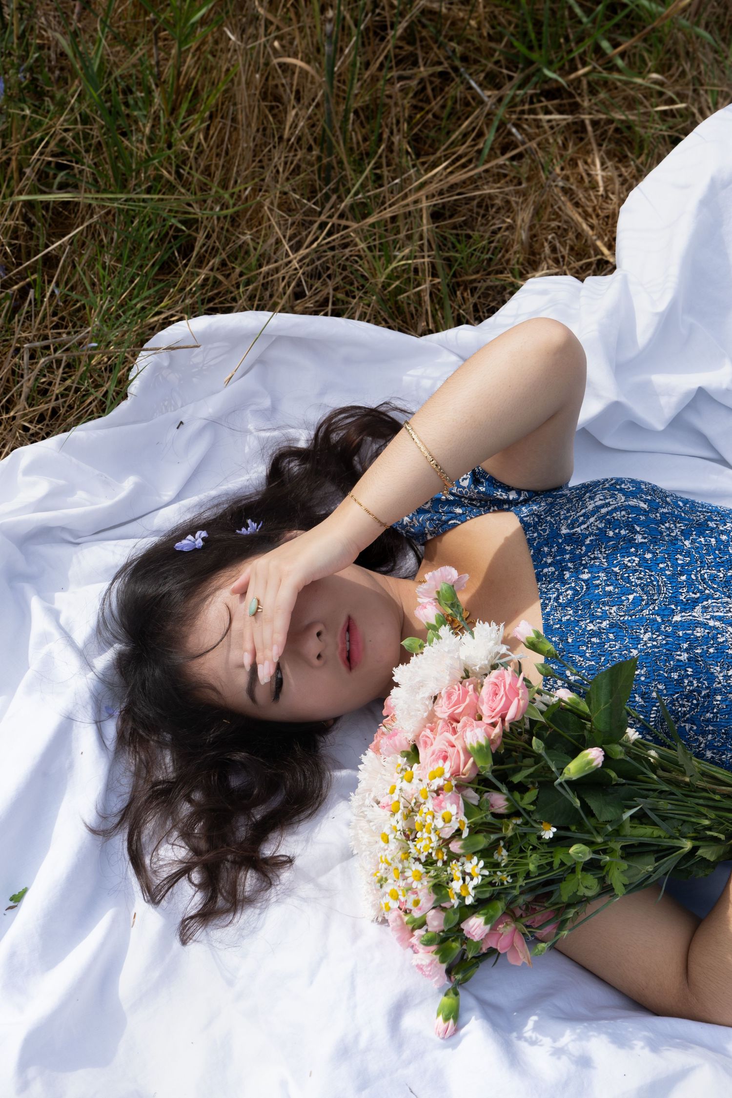 Young woman in blue floral dress lying on white blanket in grass, holding pink and white flower bouquet.