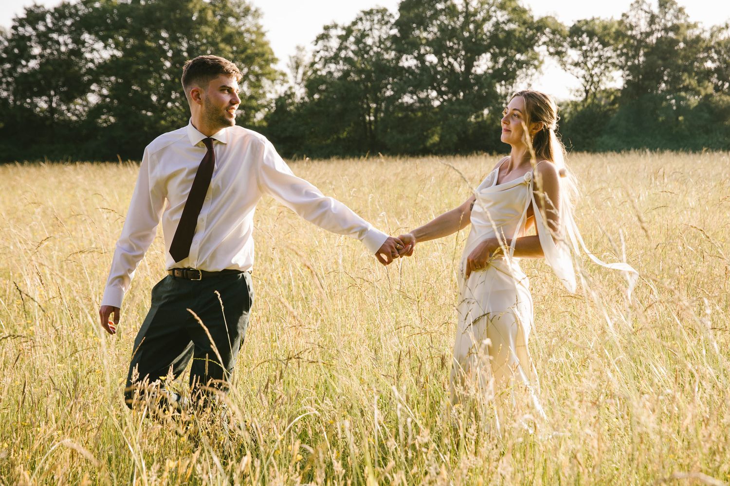 A couple holds hands walking through a golden sunlit field, man in white shirt and tie, woman in white outfit.