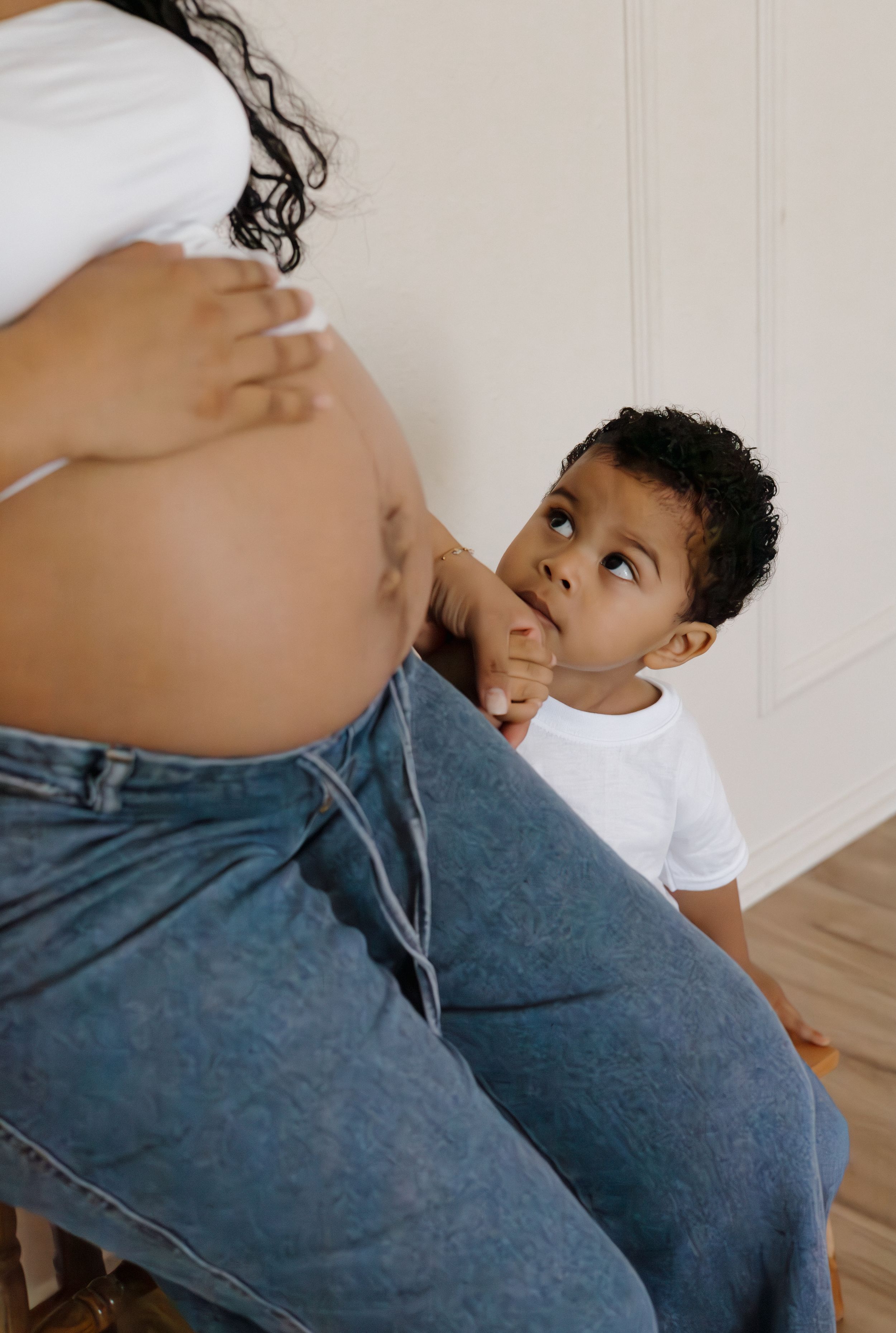 Pregnant woman in white top and jeans with toddler son touching her baby bump.