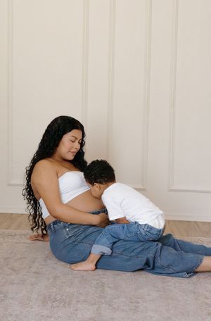 Pregnant mother with long curly hair sitting on floor as toddler son kisses her baby bump.