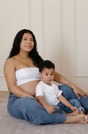 Pregnant woman in white bandeau top and jeans sitting on floor with young toddler son.