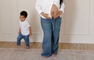 Pregnant woman in white shirt and jeans showing baby bump while toddler son walks nearby.