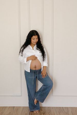 Pregnant woman with curly hair in white shirt and jeans leaning against white paneled wall.