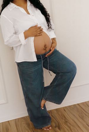Side profile of pregnant woman in white button-up shirt and wide-leg jeans cradling baby bump.