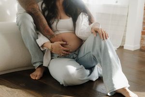 A couple sits on a hardwood floor, with a tattooed man cradling a pregnant woman's baby bump.