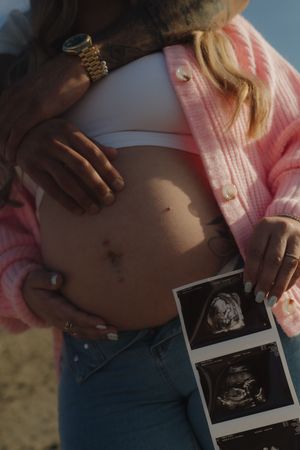 Close-up of pregnant belly with ultrasound photos held by couple on a sunny beach.