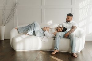 A couple relaxes on a white sofa, with a pregnant woman lying across a tattooed man's lap.