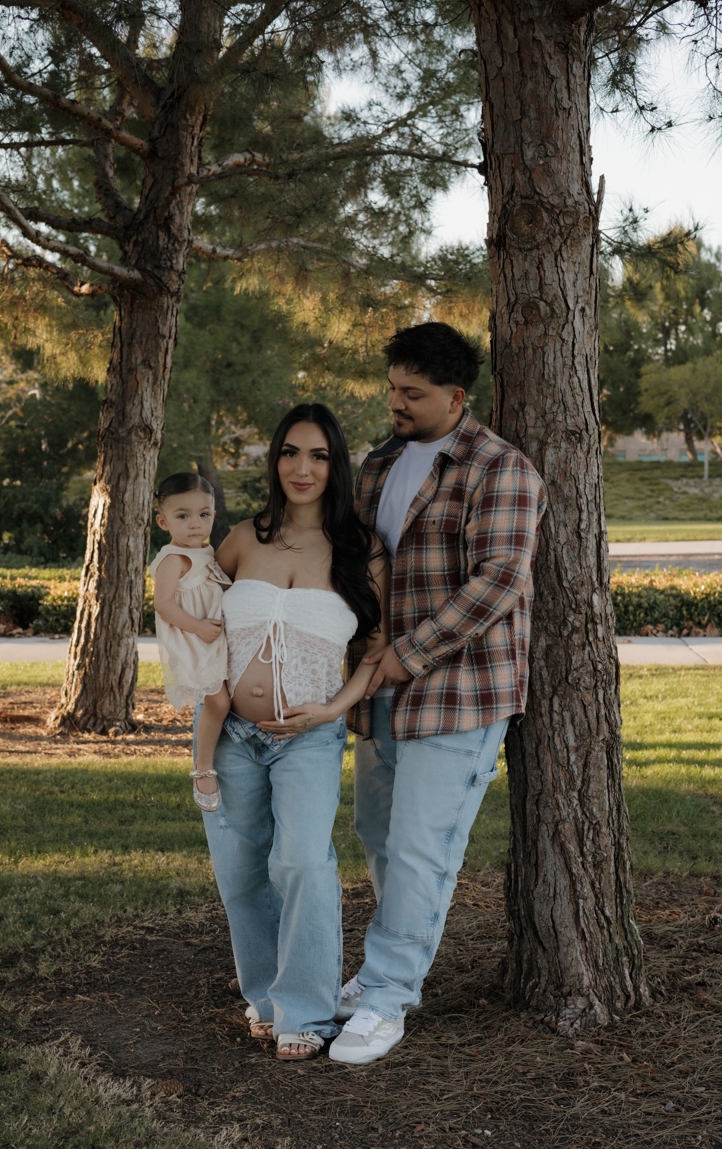 Family maternity photo with pregnant woman, partner, and toddler standing between trees outdoors.