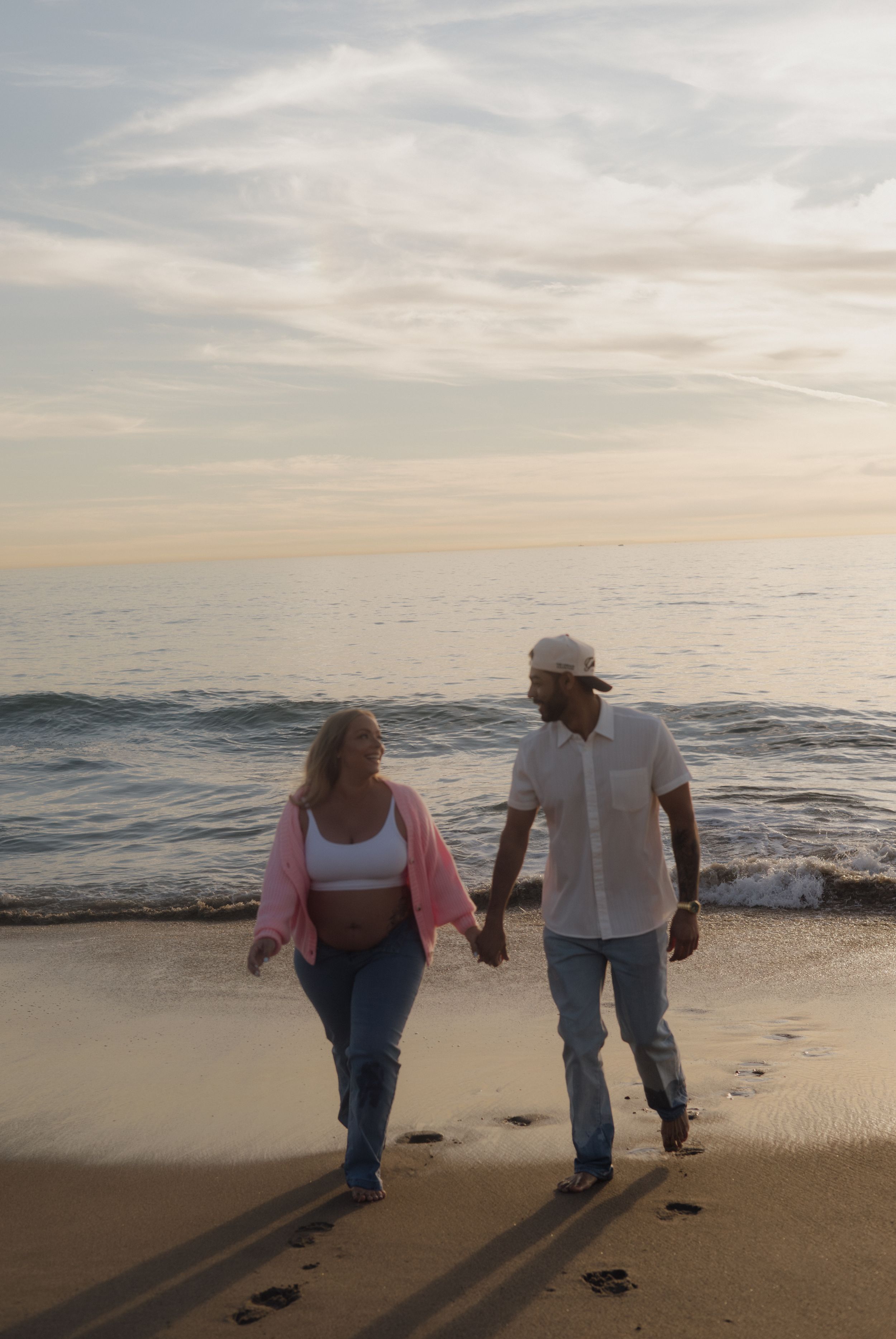 Expecting couple holding hands walking along ocean shoreline at sunset on sandy beach.