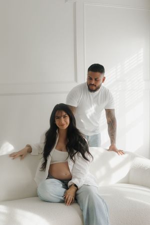 A pregnant woman in a white shirt and jeans sits on a white sofa with a tattooed man standing behind her.