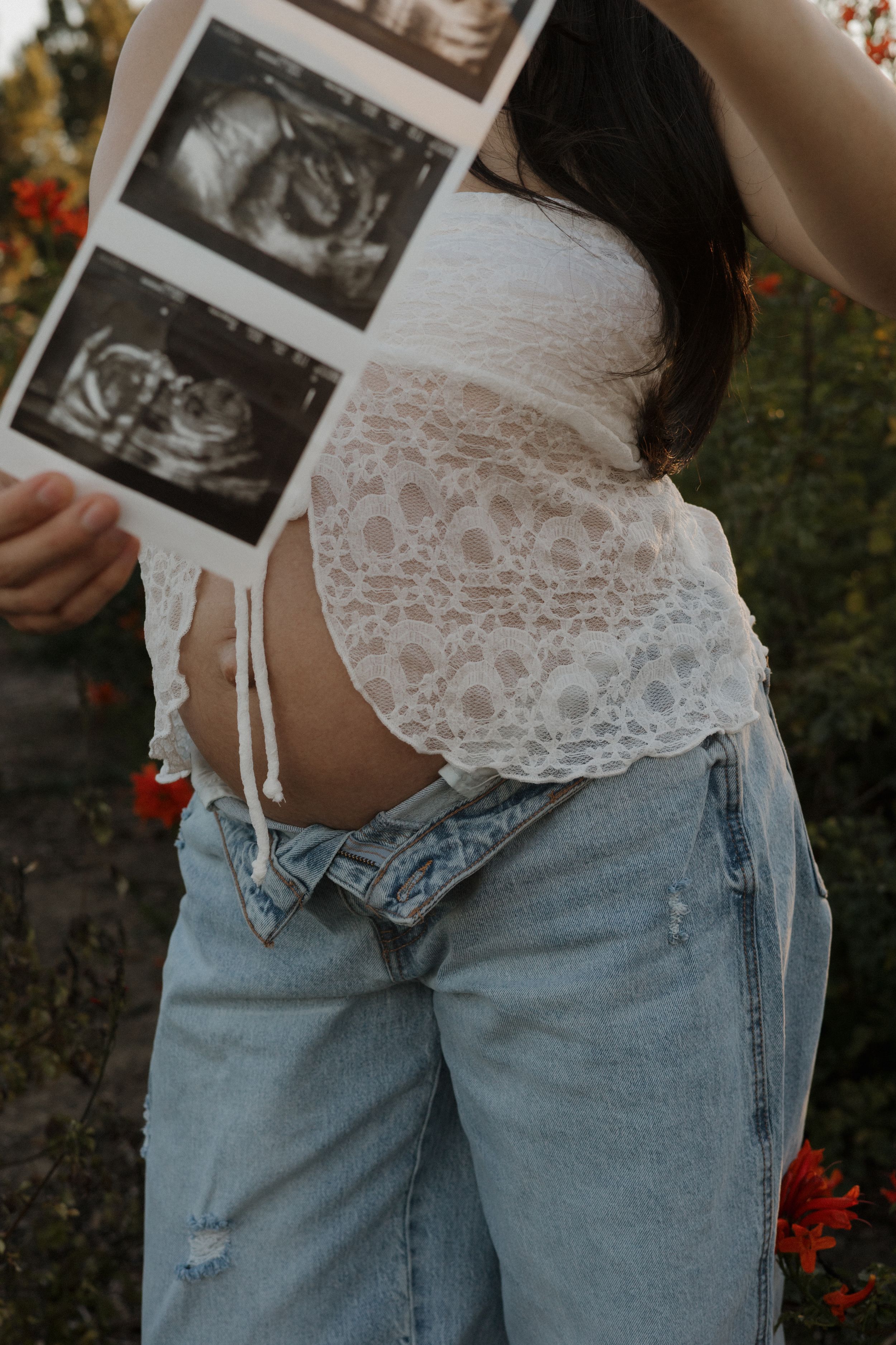 Pregnant woman displays baby ultrasound photos while showing her bare baby bump in white lace top and jeans.