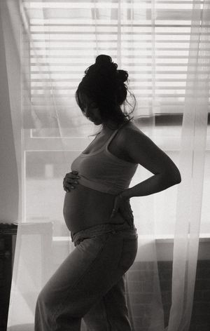 A pregnant woman in a white tank top stands silhouetted against sheer curtains in black and white.